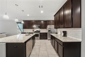 kitchen with dark brown cabinetry, appliances with stainless steel finishes, an island with sink, pendant lighting, and decorative backsplash