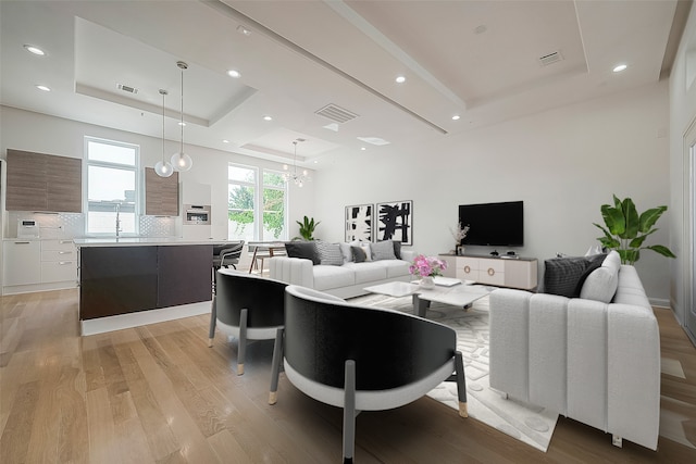 living room with sink, a tray ceiling, light hardwood / wood-style flooring, and a chandelier