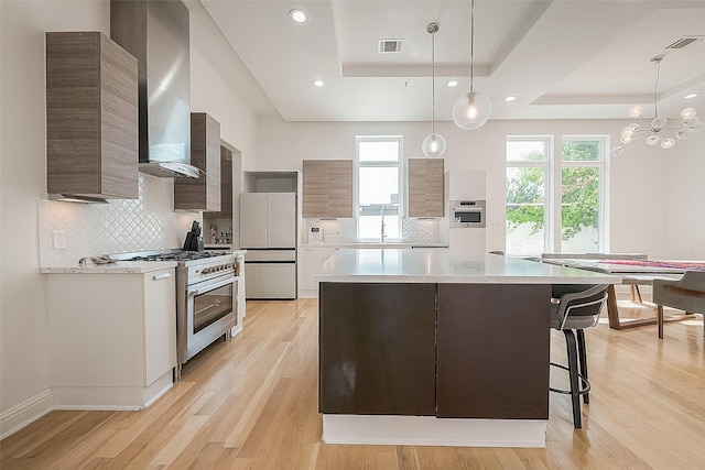 kitchen featuring pendant lighting, a tray ceiling, stainless steel appliances, wall chimney range hood, and light hardwood / wood-style flooring