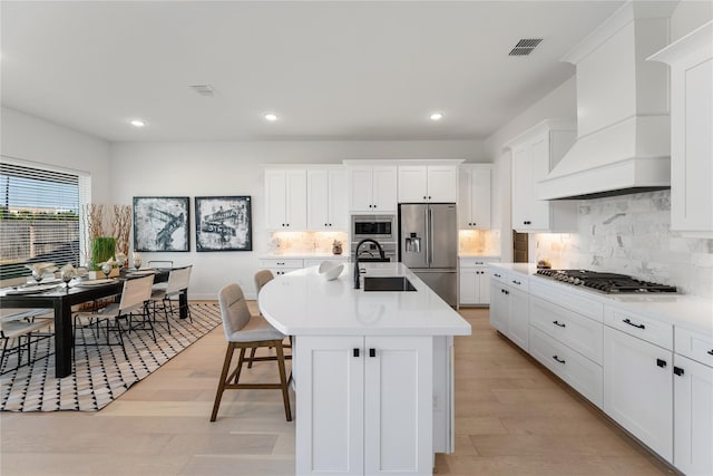 kitchen featuring a kitchen island with sink, appliances with stainless steel finishes, custom range hood, sink, and light hardwood / wood-style flooring