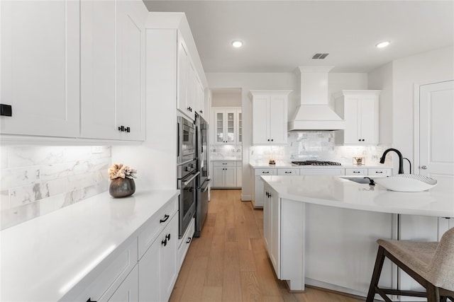 kitchen with white cabinetry, appliances with stainless steel finishes, custom range hood, light wood-type flooring, and a breakfast bar