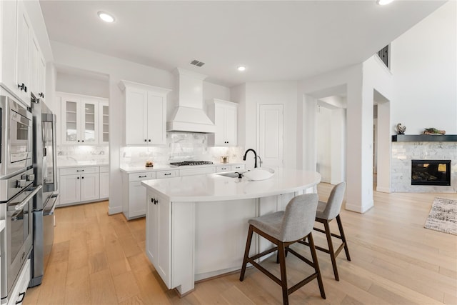 kitchen with white cabinets, light hardwood / wood-style floors, an island with sink, and premium range hood