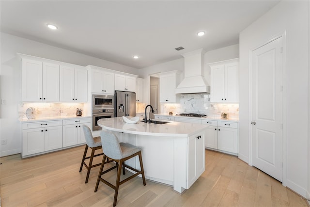 kitchen featuring sink, a kitchen island with sink, white cabinetry, appliances with stainless steel finishes, and premium range hood