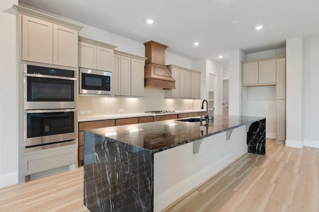 kitchen featuring custom exhaust hood, light wood-type flooring, an island with sink, and appliances with stainless steel finishes