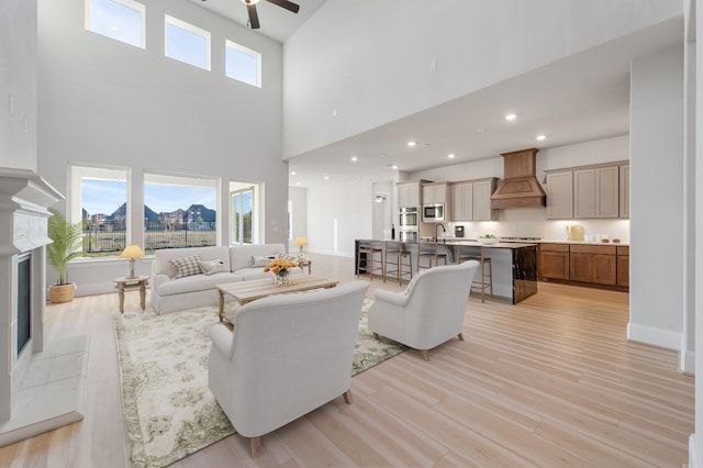 living room featuring ceiling fan, sink, a high ceiling, a premium fireplace, and light hardwood / wood-style flooring