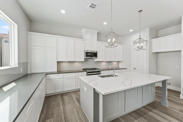 kitchen featuring appliances with stainless steel finishes, light stone counters, a center island with sink, white cabinetry, and hanging light fixtures