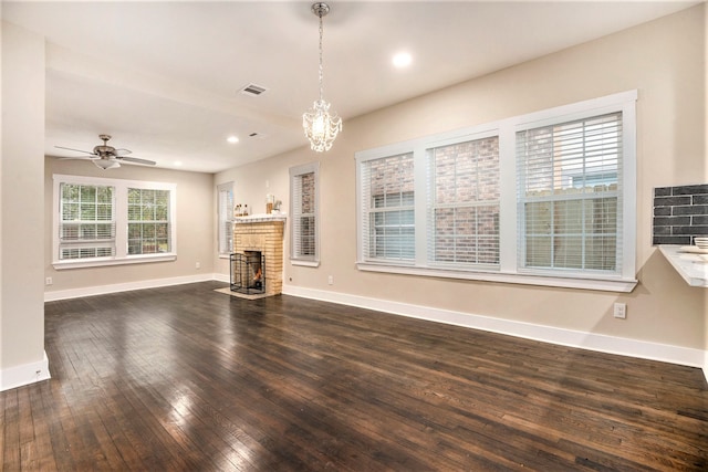 unfurnished living room with ceiling fan with notable chandelier, a brick fireplace, a wealth of natural light, and dark wood-type flooring