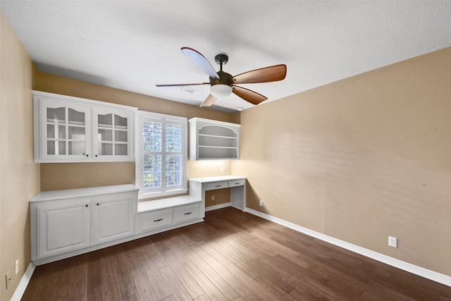 unfurnished office with ceiling fan, a textured ceiling, built in desk, and dark hardwood / wood-style floors