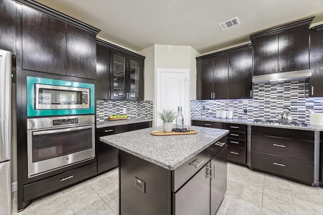 kitchen featuring tasteful backsplash, light stone counters, appliances with stainless steel finishes, dark brown cabinets, and a center island