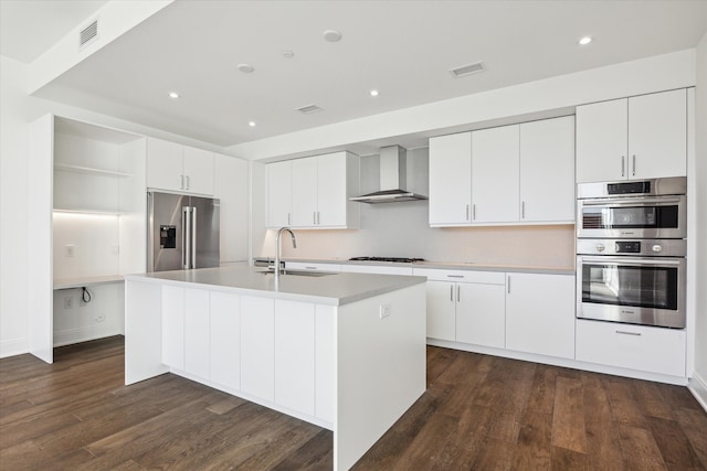 kitchen with wall chimney exhaust hood, stainless steel appliances, dark wood-type flooring, a center island with sink, and white cabinets