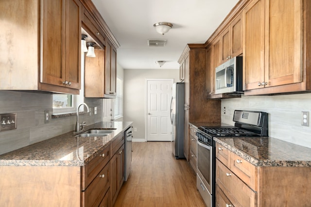 kitchen with appliances with stainless steel finishes, backsplash, sink, light hardwood / wood-style flooring, and dark stone countertops