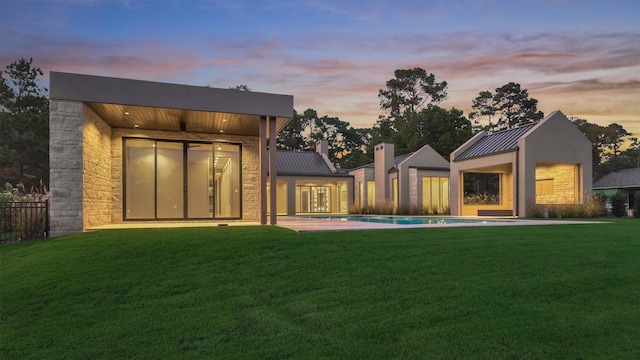 back of house at dusk featuring metal roof, a standing seam roof, stone siding, and an outdoor pool