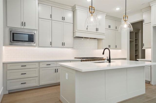 kitchen featuring an island with sink, a sink, stainless steel microwave, decorative backsplash, and light wood-style flooring