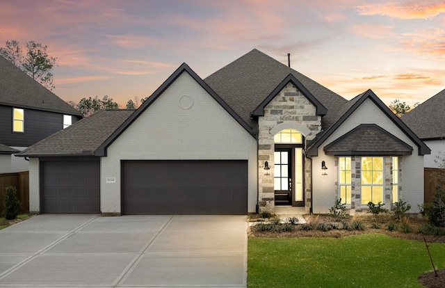 french country home featuring an attached garage, stone siding, a shingled roof, and concrete driveway