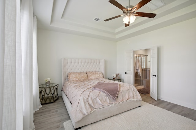 bedroom featuring light wood-type flooring, a tray ceiling, ensuite bath, and ceiling fan