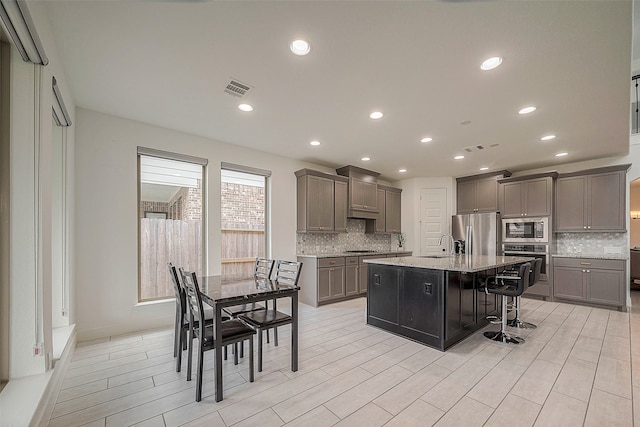 kitchen with tasteful backsplash, stainless steel appliances, a kitchen bar, light stone countertops, and an island with sink