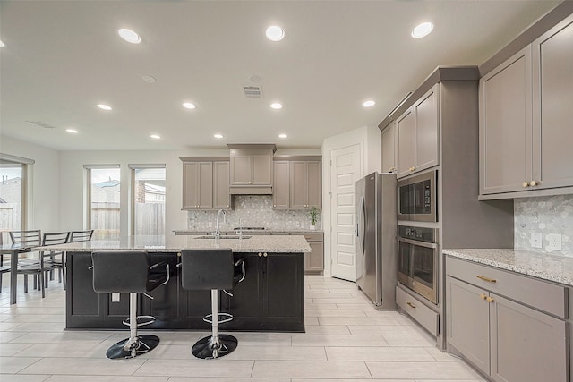 kitchen featuring gray cabinetry, appliances with stainless steel finishes, light stone counters, and a kitchen island with sink