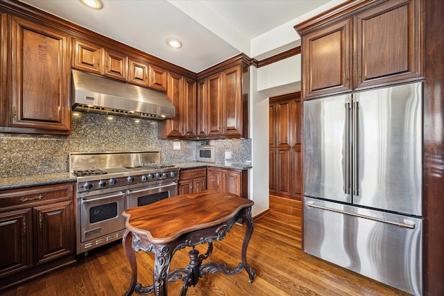 kitchen with dark hardwood / wood-style floors, dark stone countertops, range hood, and appliances with stainless steel finishes