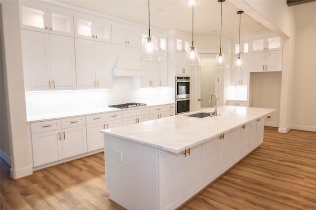 kitchen featuring white cabinetry, sink, light stone countertops, premium range hood, and an island with sink