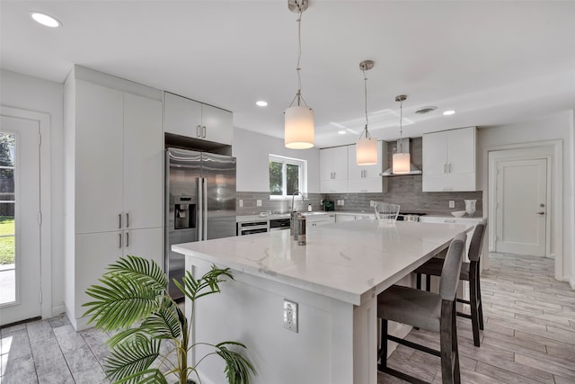 kitchen with hanging light fixtures, wall chimney range hood, high end refrigerator, light hardwood / wood-style flooring, and white cabinets