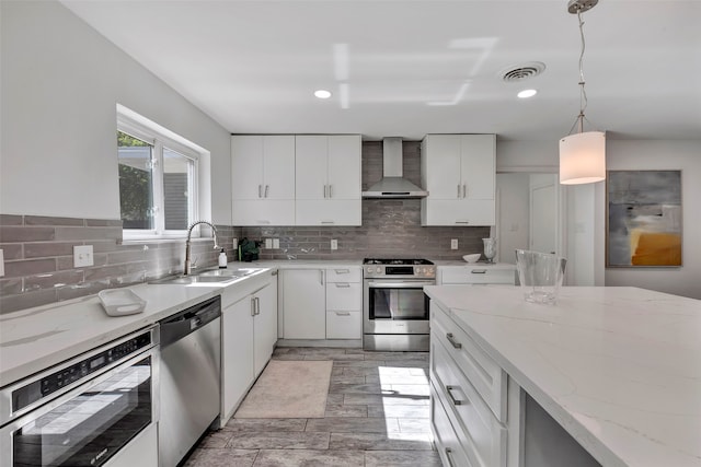 kitchen with wall chimney range hood, sink, decorative light fixtures, white cabinetry, and stainless steel appliances