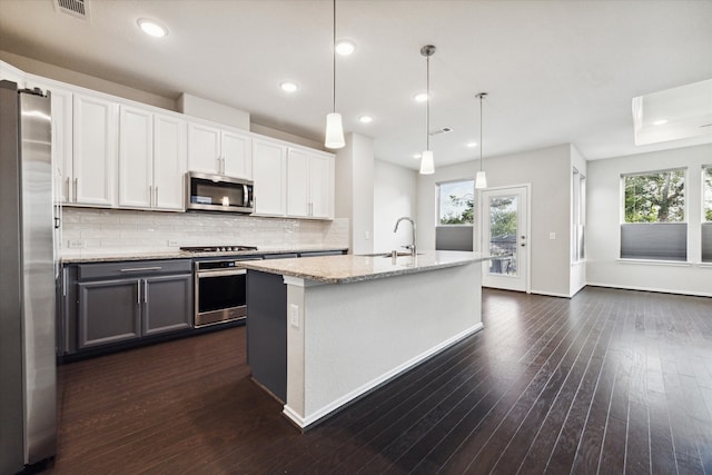 kitchen with appliances with stainless steel finishes, hanging light fixtures, white cabinets, dark wood-type flooring, and a kitchen island with sink