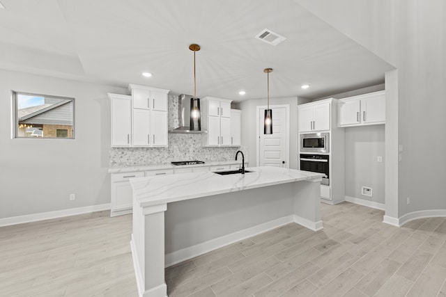 kitchen featuring a center island with sink, wall chimney range hood, light stone countertops, pendant lighting, and appliances with stainless steel finishes