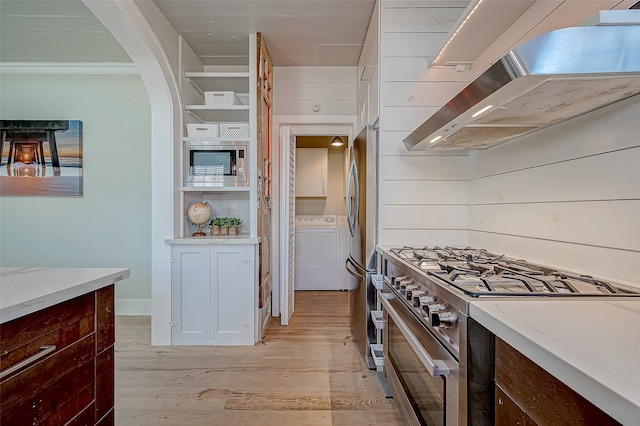 kitchen featuring light stone counters, exhaust hood, washer and clothes dryer, light wood-type flooring, and appliances with stainless steel finishes