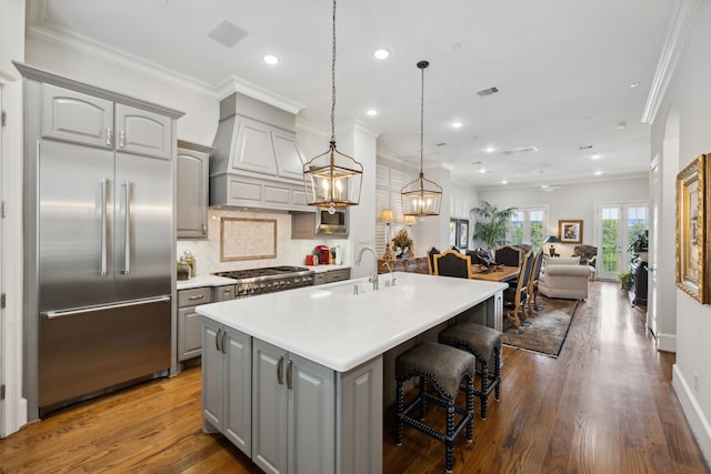 kitchen with sink, gray cabinetry, built in refrigerator, hanging light fixtures, and a center island with sink