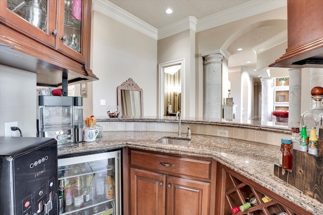 kitchen with light stone counters, sink, decorative columns, ornamental molding, and wine cooler