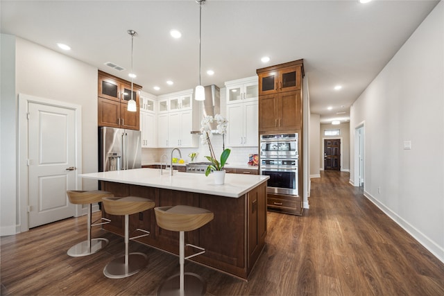 kitchen featuring wall chimney exhaust hood, an island with sink, backsplash, pendant lighting, and appliances with stainless steel finishes