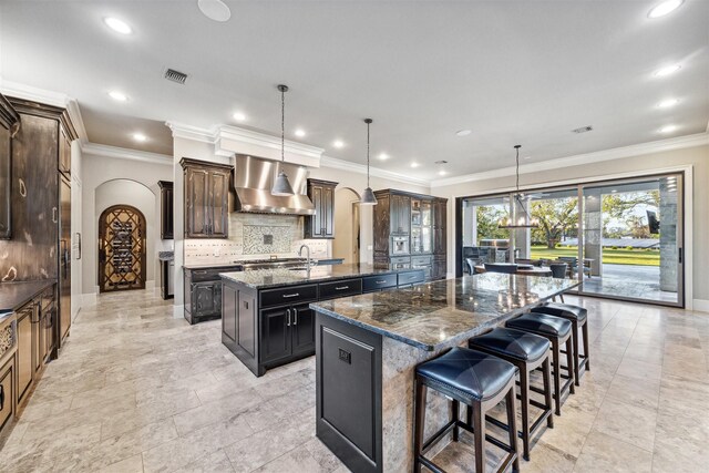 kitchen with dark brown cabinetry, a large island, wall chimney range hood, dark stone counters, and pendant lighting