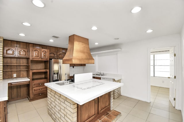 kitchen featuring stainless steel refrigerator with ice dispenser, premium range hood, a kitchen island with sink, and sink