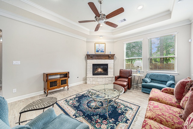 living room with a raised ceiling, ceiling fan, crown molding, light hardwood / wood-style flooring, and a stone fireplace