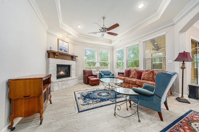 living room featuring a stone fireplace, light hardwood / wood-style flooring, ceiling fan, ornamental molding, and a tray ceiling