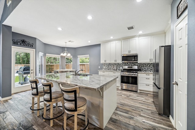 kitchen with white cabinetry, appliances with stainless steel finishes, decorative light fixtures, a kitchen island with sink, and light stone counters