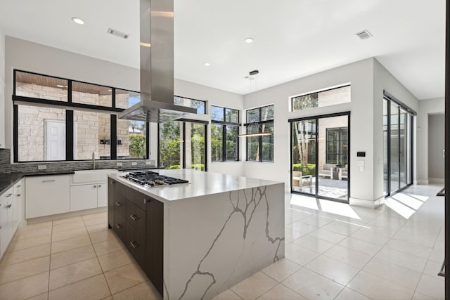 kitchen with backsplash, light stone counters, island range hood, white cabinetry, and stainless steel gas stovetop