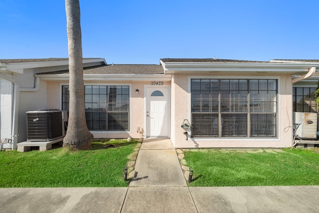 view of front facade with a front yard, a shingled roof, cooling unit, and stucco siding