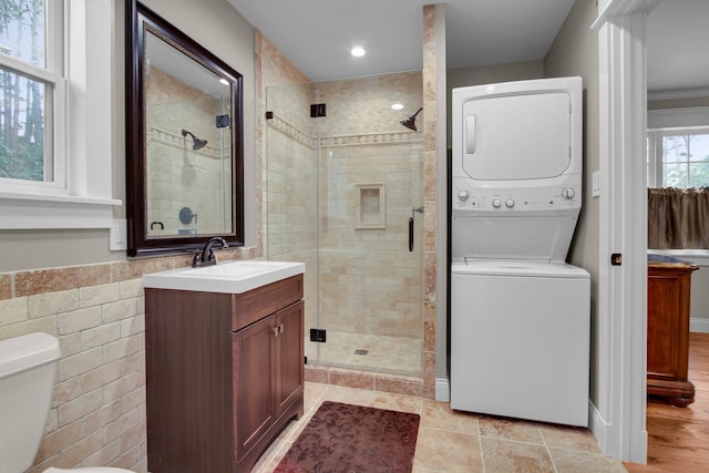 bathroom featuring stacked washer / dryer, toilet, vanity, a shower with shower door, and tile walls