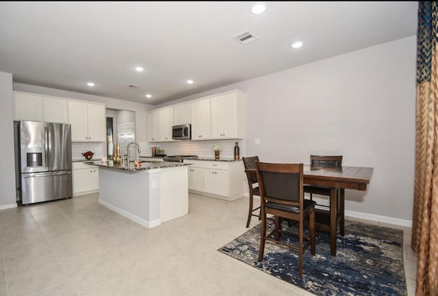 kitchen with white cabinets, stainless steel appliances, a kitchen island with sink, and dark stone counters