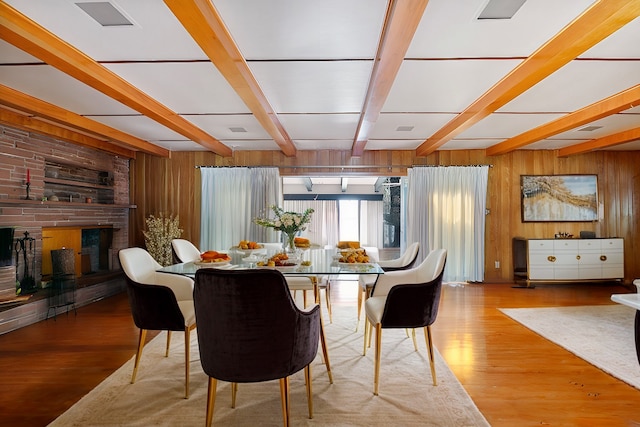 dining room featuring beam ceiling, a brick fireplace, wooden walls, and light wood-type flooring