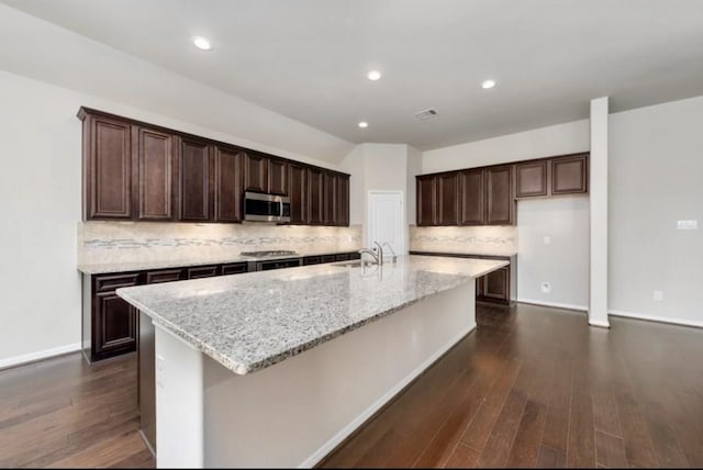 kitchen featuring tasteful backsplash, light stone countertops, an island with sink, and dark wood-type flooring