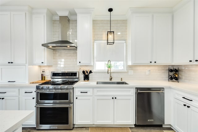 kitchen featuring wall chimney range hood, sink, decorative light fixtures, white cabinetry, and stainless steel appliances