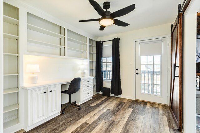 unfurnished office featuring ceiling fan, a barn door, built in desk, and dark wood-type flooring