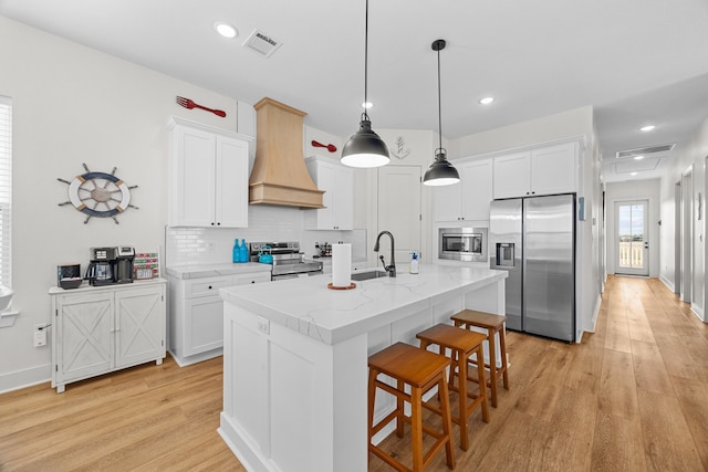 kitchen with stainless steel appliances, light hardwood / wood-style floors, custom range hood, white cabinets, and a center island with sink