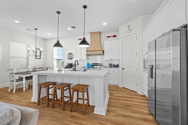 kitchen featuring a center island with sink, appliances with stainless steel finishes, custom range hood, light hardwood / wood-style floors, and white cabinets