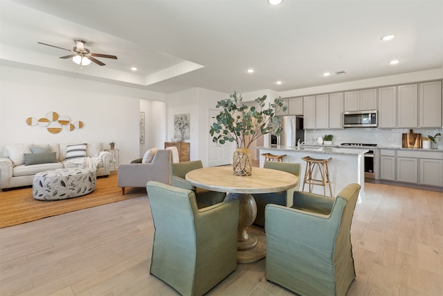 dining area featuring a raised ceiling, ceiling fan, sink, and light wood-type flooring