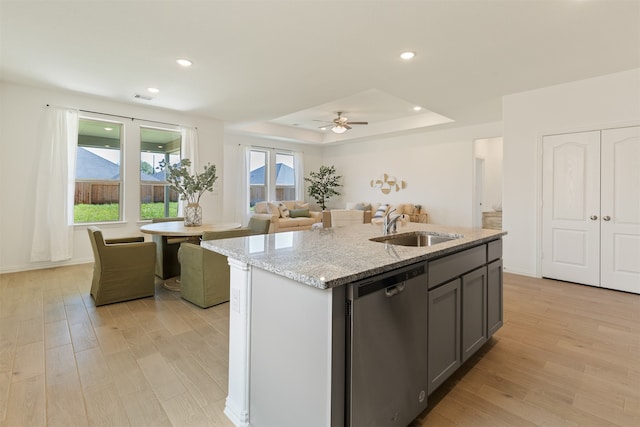 kitchen with dishwasher, sink, a raised ceiling, light stone counters, and an island with sink