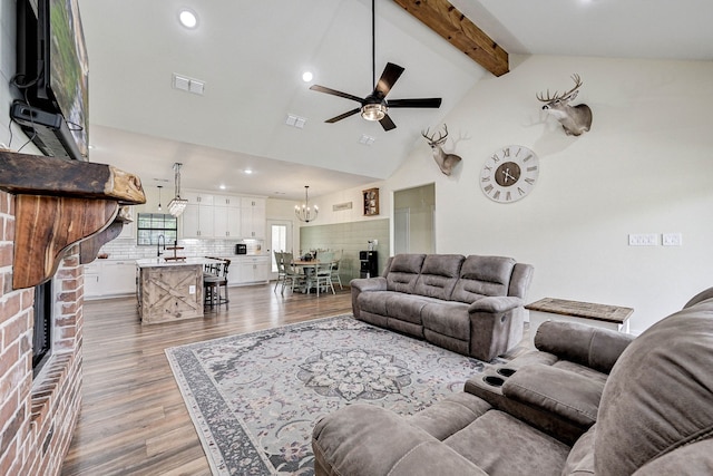 living room featuring lofted ceiling with beams, ceiling fan with notable chandelier, sink, a brick fireplace, and hardwood / wood-style flooring