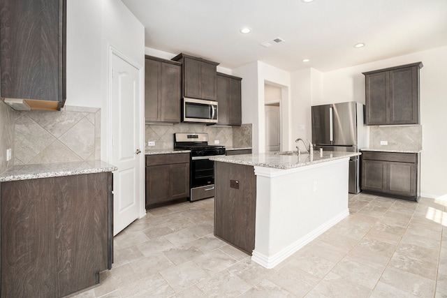 kitchen featuring light stone countertops, a kitchen island with sink, stainless steel appliances, decorative backsplash, and dark brown cabinets